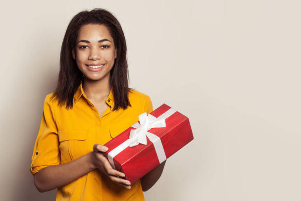 Happy pretty african american woman holding gift and looking at the camera against white wall background - 写真・画像