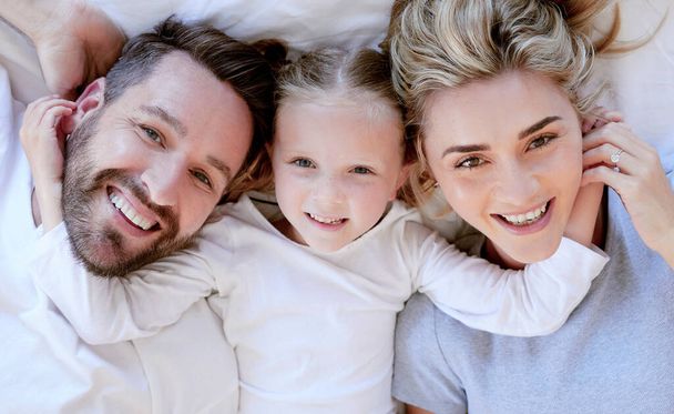 Portrait of happy carefree caucasian family in pyjamas from above lying cosy together in bed at home. Loving parents with little daughter. Cute young girl touching and hugging her mom and dads face. - Foto, afbeelding