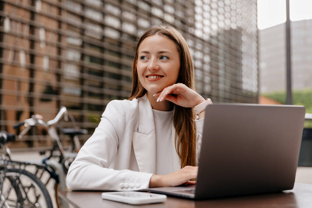 Close up portret uśmiechniętej stylowej Europejki w białej kurtce siedzi na ulicy kawiarni i pracuje na laptopie. Atrakcyjny biznes lady w kurtce spoczywa na zewnątrz.  - Zdjęcie, obraz