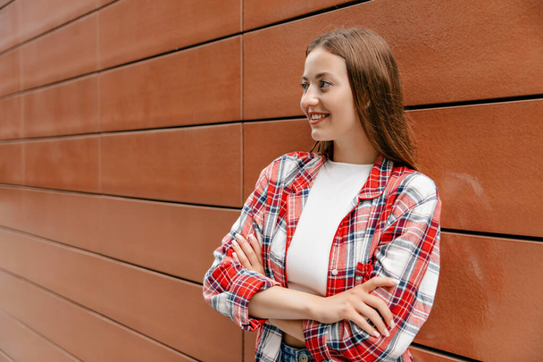 Outdoor portrait of happy woman with long hair wearing white top and checkered shirt posing with crossed arms and wonderful smile on background of brown wall  - Foto, afbeelding