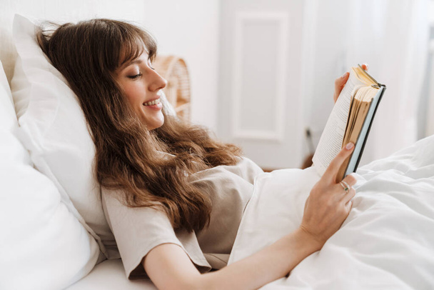 White young woman smiling and reading book while lying in bed at home - Foto, immagini