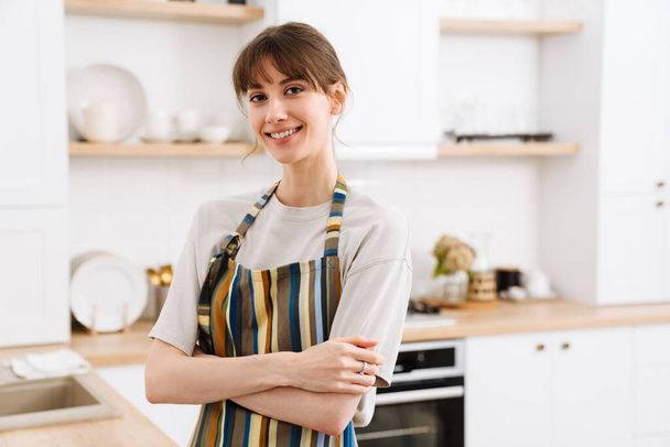 White young woman wearing apron smiling while standing in kitchen at home - Foto, immagini