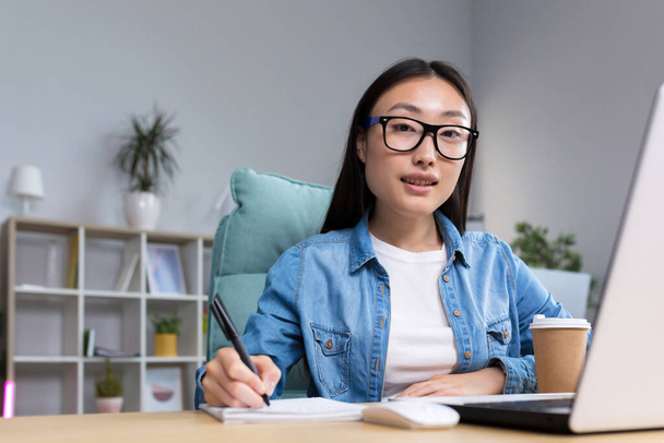 Job interview. A young beautiful Asian business woman in glasses sits at a desk, conducts an interview in the office, hires, writes down a questionnaire in a notebook, interviews the interlocutor - Foto, imagen