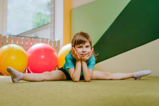Little girl sitting on the twine looking at camera in sport gym. Preschooler doing exercises on the mat. Health concept. - Foto, immagini