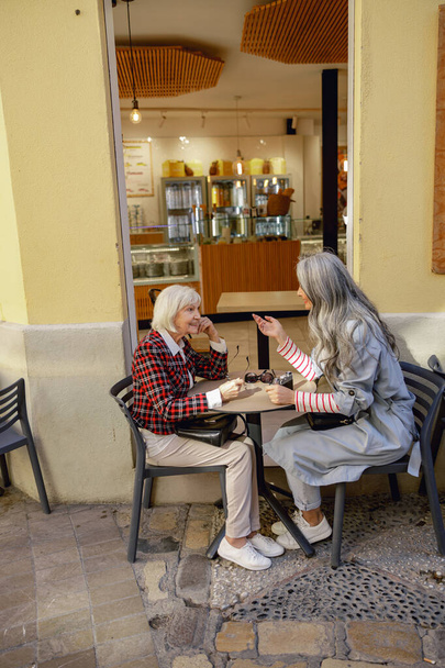 Yaşlanmış bayan arkadaşlar günlük giysiler giyip sokak kafesinde masada otururken sohbet ediyorlar. - Fotoğraf, Görsel