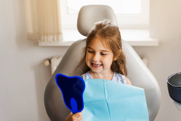 Child looking in the mirror at the dentist. Happy child patient of dentistry. Teeth treatment. Attractive kid girl sitting in dental office and smiling - Photo, Image