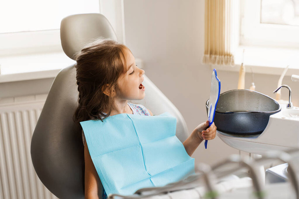 Child looking in the mirror at the dentist. Happy child patient of dentistry. Teeth treatment. Attractive kid girl sitting in dental office and smiling - Φωτογραφία, εικόνα