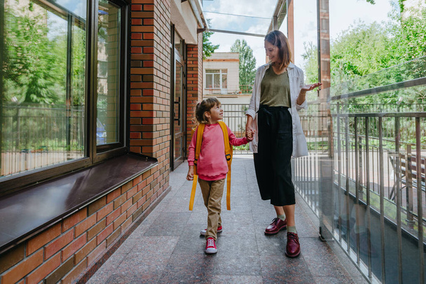 Mother leading her daughter at school. Young woman and little 6 years old girl near elementary school building. Back to school concept. - Фото, зображення
