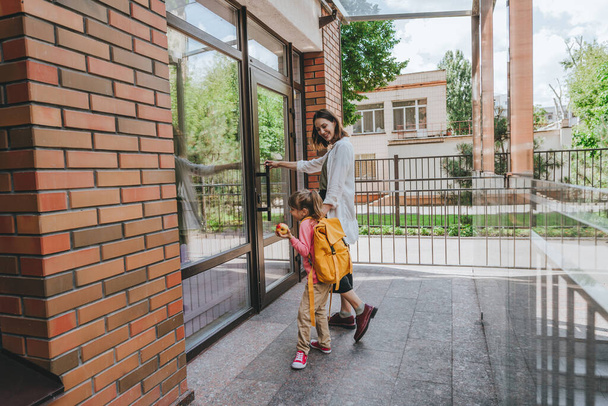 Mother leading her daughter at school. Young woman and little 6 years old girl near elementary school building opening the door. Back to school concept. - Photo, image