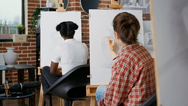 Mujer joven usando lápiz para dibujar jarrón sobre lienzo blanco, aprendiendo nuevas habilidades artísticas en clase de arte. Estudiante dibujo inspiración modelo en lección práctica con multiétnico grupo de personas. - Foto, imagen
