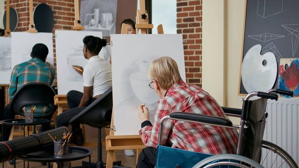 Старшая женщина в инвалидной коляске рисует вазу с карандашом на холсте и мольберте, посещает художественные классы со студентами и преподавателем. Люди с ограниченными возможностями учатся рисовать для роста. - Фото, изображение