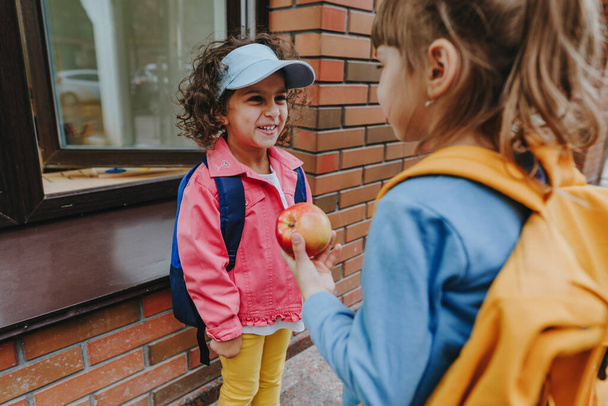 Back view of two little girls with backpacks going to an elementary school. Friend giving an apple. Back to school concept. Selective focus on girls. - Фото, изображение