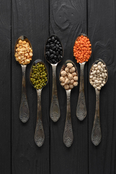Variety of legumes in old silver spoons on a black wooden background. - Foto, Imagem