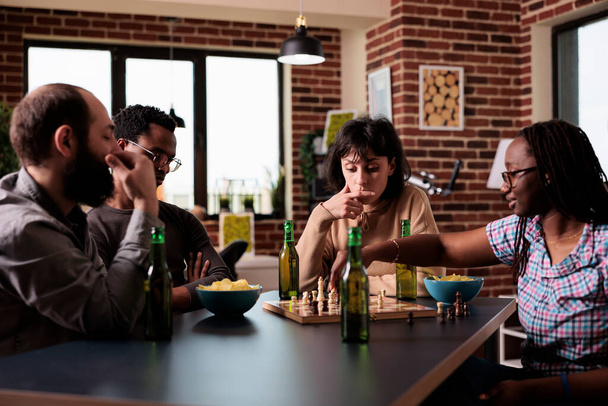 Intelligente multiethnische Gruppe von Menschen, die am Tisch sitzen, während sie Schach spielen. Glückliche Freunde sitzen zu Hause im Wohnzimmer zusammen und genießen Strategiespiele bei Getränken und Snacks.. - Foto, Bild