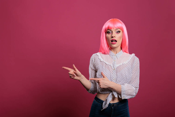 Attractive young woman with pink hair pointing sideways in studio, indicating left or right directions in front of camera. Recommending that way with raised arms and index fingers. - 写真・画像