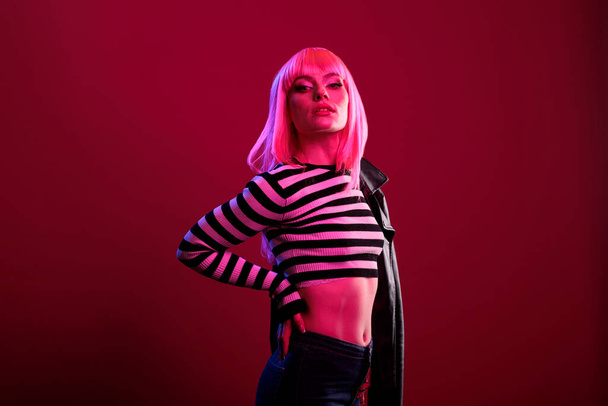 Portrait of confident model with leather jacket posing in dark pink light, using fluorescent lighting in studio to take pictures. Cool stylish rocker with funky hairstyle and punk clothes. - Photo, Image