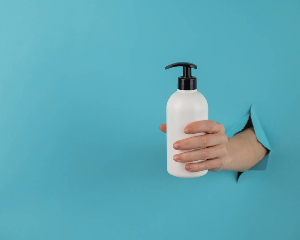 A womans hand sticks out of a hole in a blue paper background and holds a skin lotion pump - Foto, Imagem