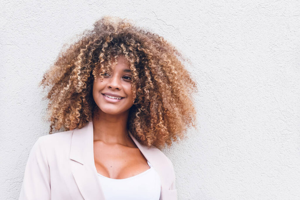 face of beautiful afro woman with curly hair on white wall background in the city smiling happy - 写真・画像