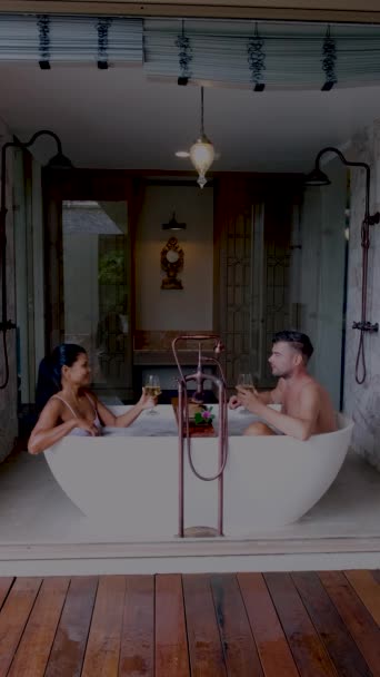 Donne asiatiche e uomini europei si rilassano nelle vasche da bagno durante le vacanze, una vacanza di lusso. Coppia uomo e donne che bevono vino nella vasca da bagno - Filmati, video