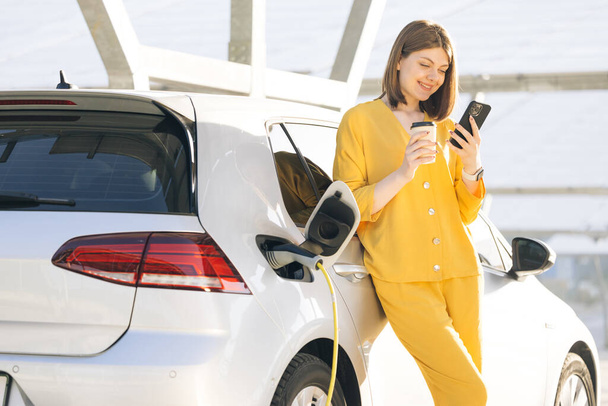 Caucasian woman using smart phone and waiting power supply connect to electric vehicles for charging the battery in car. Plug charging an Electric car from charging station. - Photo, Image