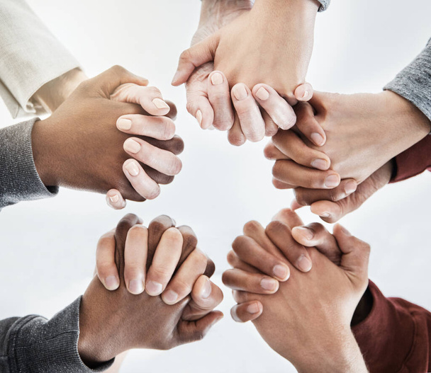 Below close up view of people holding hands in circle shape. A group of people putting their hands together while standing in a huddle inside against a clear grey background. Anything is possible with - Photo, Image