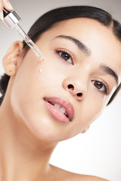 Portrait of a young beautiful mixed race woman applying oil to her face while standing against a grey studio background alone. One hispanic female doing her skincare routine while standing against a - Foto, imagen