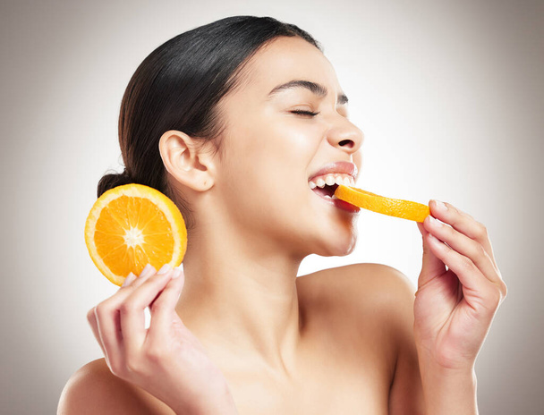 Young happy beautiful mixed race woman eating an orange while posing against a grey background. One cheerful hispanic female eating a healthy fruit against a background. - Fotografie, Obrázek