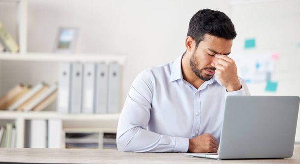 Young mixed race businessman suffering from a headache working on a laptop in an office alone at work. One hispanic man stressing while working. Male boss feeling anxious sitting at a desk. - Foto, imagen