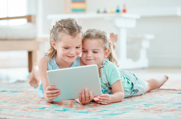 zwei entzückende Mädchen, die ein digitales Tablet benutzen, während sie zu Hause auf dem Boden liegen. - Foto, Bild