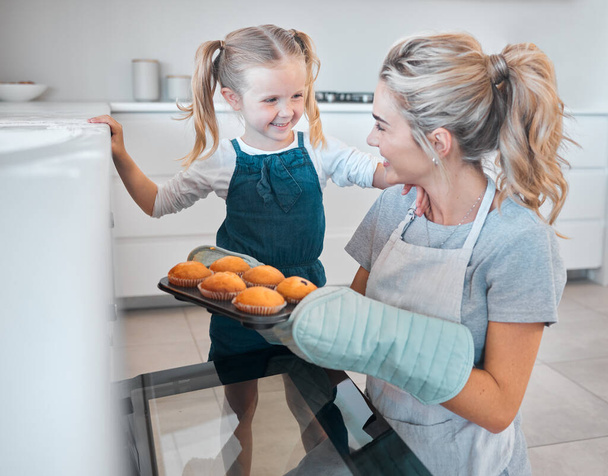 Mother and daughter removing tray of muffins from the oven. Happy woman baking muffins with her daughter. Little girl bonding with her mother and baking. Parent taking muffins from the oven. - Photo, Image