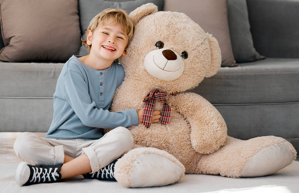 Portrait of one happy little caucasian boy smiling while hugging a big and cosy fluffy teddy bear on the floor in the lounge at home. Adorable kid relaxing and playing with soft stuffed toy alone. - Фото, зображення
