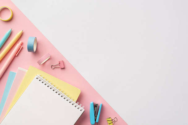 School accessories concept. Top view photo of colorful stationery copybooks pens adhesive tape binder clips and stapler on bicolor white and pink background with empty space - Photo, Image
