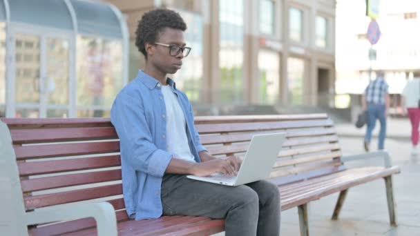 Afrikaner mit Laptop lächelt in Kamera, während er draußen auf Bank sitzt - Filmmaterial, Video