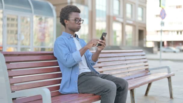 African Man Celebrating Online Success on Smartphone while Sitting Outdoor on Bench - Metraje, vídeo