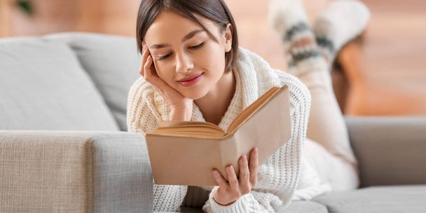 Piękna młoda kobieta czyta książkę leżąc na kanapie w domu - Zdjęcie, obraz