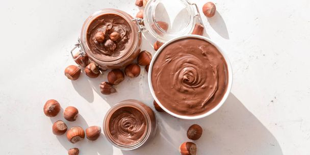 Jars and bowl with tasty chocolate paste and hazelnuts on light background, top view - Photo, image