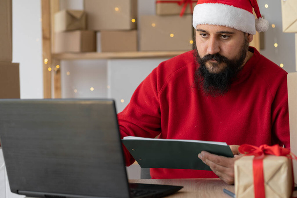 Happy Indian Santa Claus in hat using laptop while sitting at home table in workshop on Christmas Eve. online shopping sales on christmas holiday e-commerce website concept - Fotoğraf, Görsel
