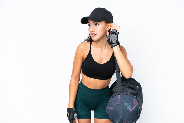 Young sport woman with sport bag isolated on white background listening to something by putting hand on the ear - Fotoğraf, Görsel