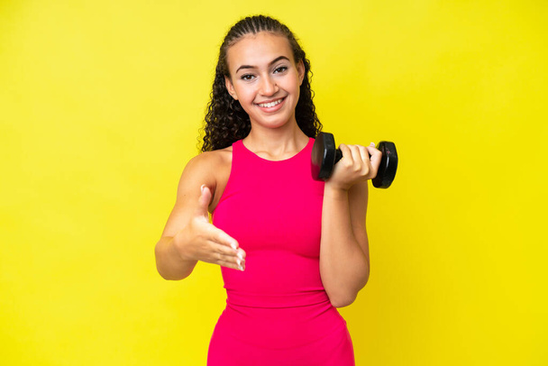 Jonge sport vrouw maken gewichtheffen geïsoleerd op gele achtergrond schudden handen voor het sluiten van een goede deal - Foto, afbeelding