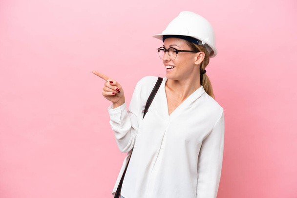 Young architect Russian woman with helmet and holding blueprints isolated on pink background pointing finger to the side and presenting a product - Photo, Image
