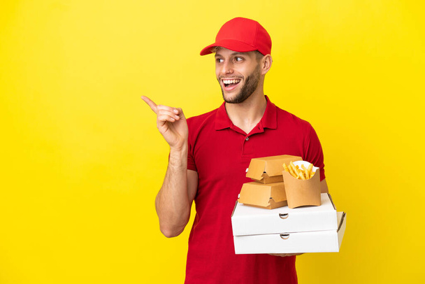 Pizzalieferant nimmt Pizzakartons und Burger über isoliertem Hintergrund in die Hand und hebt den Finger - Foto, Bild