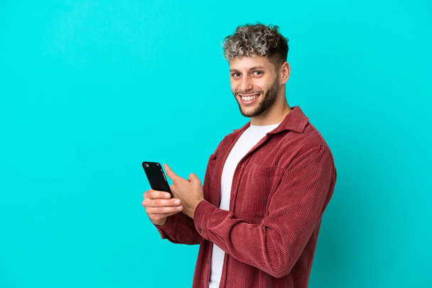 Young handsome caucasian man isolated on blue background using mobile phone and pointing back - Foto, afbeelding