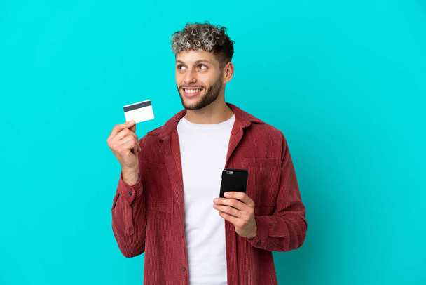Молодий красивий кавказький чоловік ізольований на синьому фоні, купуючи з мобільним телефоном з кредитною карткою під час мислення
 - Фото, зображення