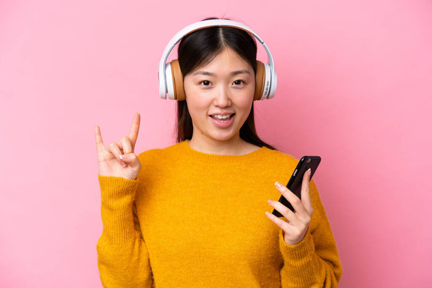 Junge Chinesin isoliert auf rosa Hintergrund Musik hören mit einem Handy machen Rock-Geste - Foto, Bild