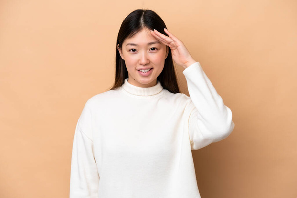 Young Chinese woman isolated on beige background saluting with hand with happy expression - Фото, изображение