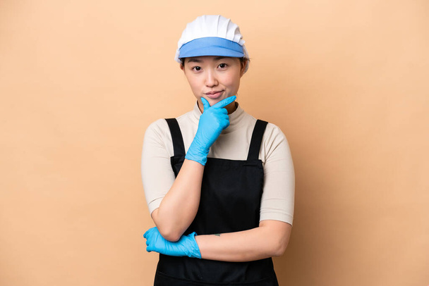 Young Chinese Fishmonger woman wearing an apron and holding a raw fish isolated on pink background thinking - Foto, Bild
