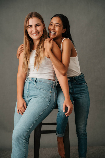 Close up of two female friends embracing and smiling and sharing a stool in the studio . High quality photo - Foto, immagini