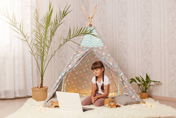 Horizontal shot of charming little girl sitting in wigwam and watching cartoons on laptop, having positive facial expression, looking at notebook screen, enjoying interesting video. - Φωτογραφία, εικόνα