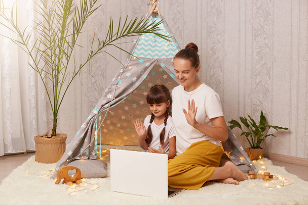 Horizontal shot of dark haired Caucasian woman and her daughter wearing white t shirts sitting in wigwam in front of laptop, waving hands, saying hello, smiling, having positive faces. - Photo, Image