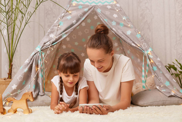 Horizontal shot of woman and her daughter laying in peetee tent and watching cartoons on mobile phone, spending time together at home, having fun in wigwam. - 写真・画像